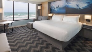 A hotel room with a large bed, a desk, and a chair. There are two wall-mounted lamps and a large window with a view of the ocean. - Hilton Cocoa Beach Oceanfront (Cocoa Beach)