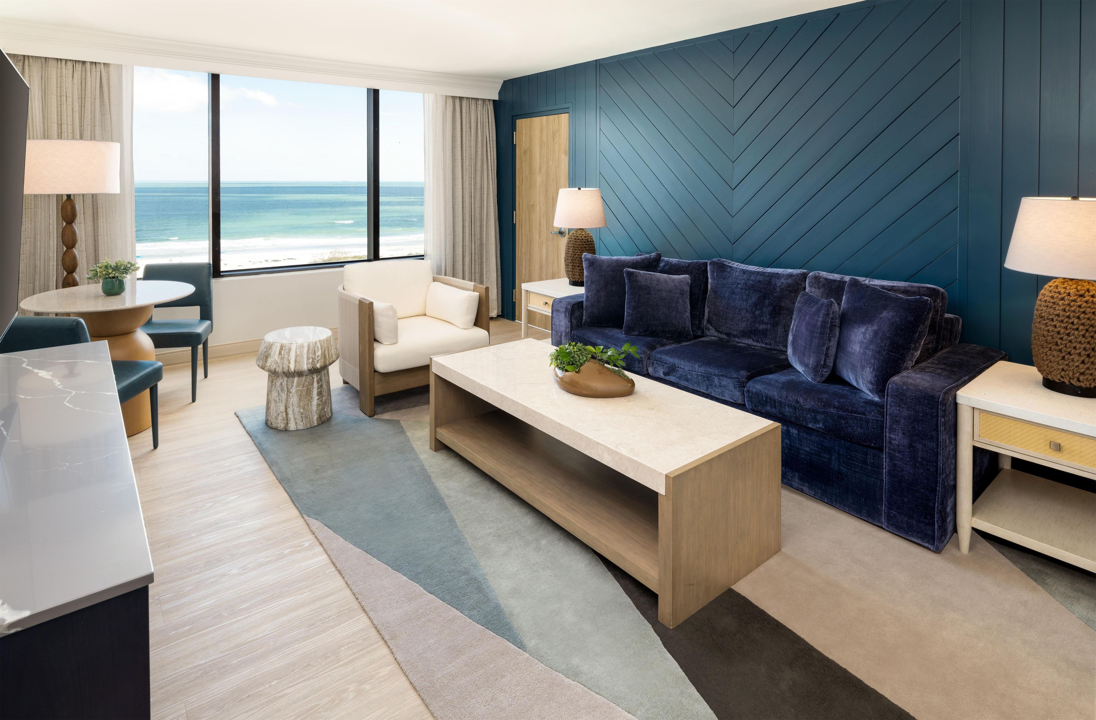 A modern hotel room with a large window offering a beach view, a blue accent wall, a sofa, a coffee table, and a dining area with a table and chairs.