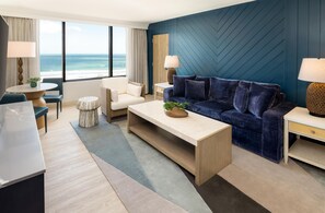 Habitación de hotel moderna con un ventanal que ofrece vistas a la playa, una pared azul de acento, un sofá, una mesa de centro y un comedor con mesa y sillas.