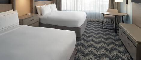 Ein Hotelzimmer mit zwei Betten, einem Schreibtisch, einem Stuhl, einem Gemälde und einem großen Fenster mit Vorhängen.
