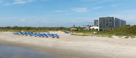 A beach with blue umbrellas and lounge chairs, a building with multiple floors, and a clear sky.