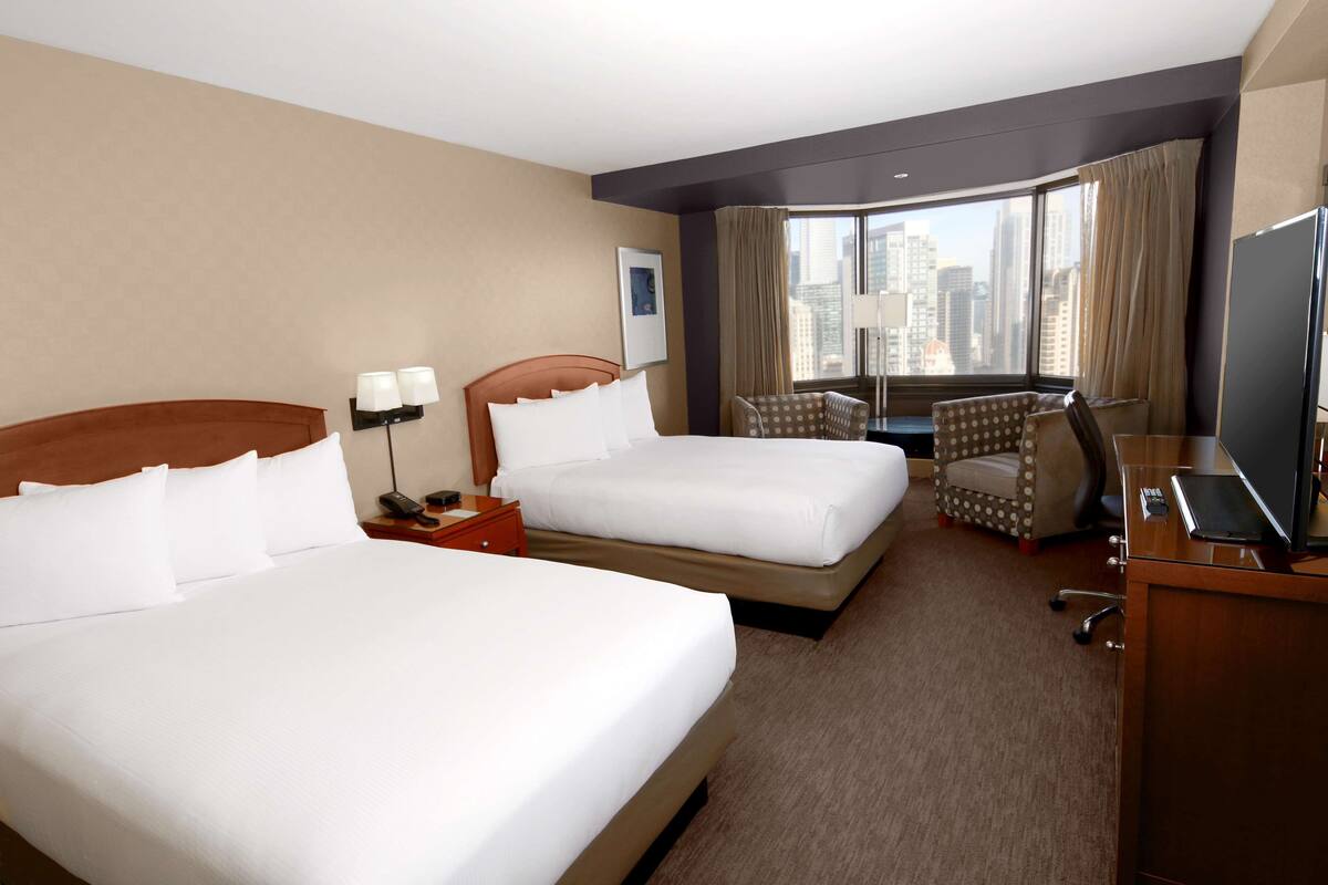 A hotel room with two beds, a desk, a chair, and a large window with a city view.