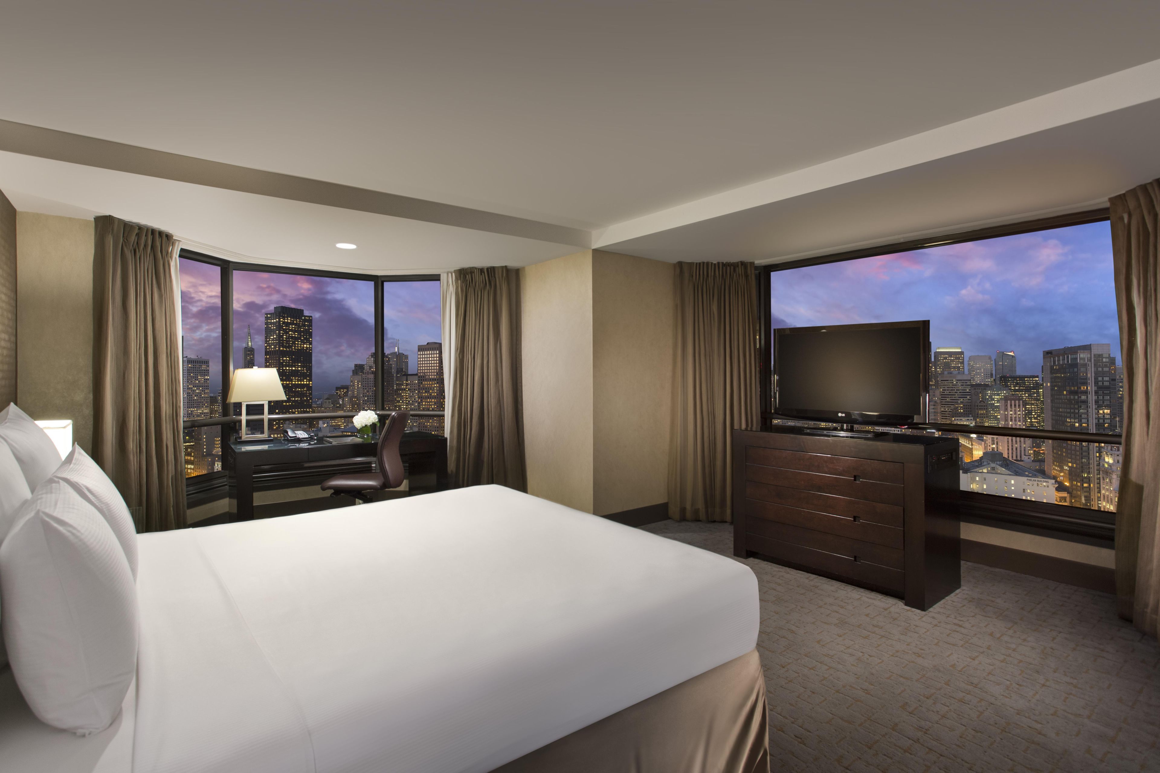 Habitación de hotel con una cama grande, un escritorio con lámpara, cómoda y televisor. La habitación tiene vista a la ciudad.