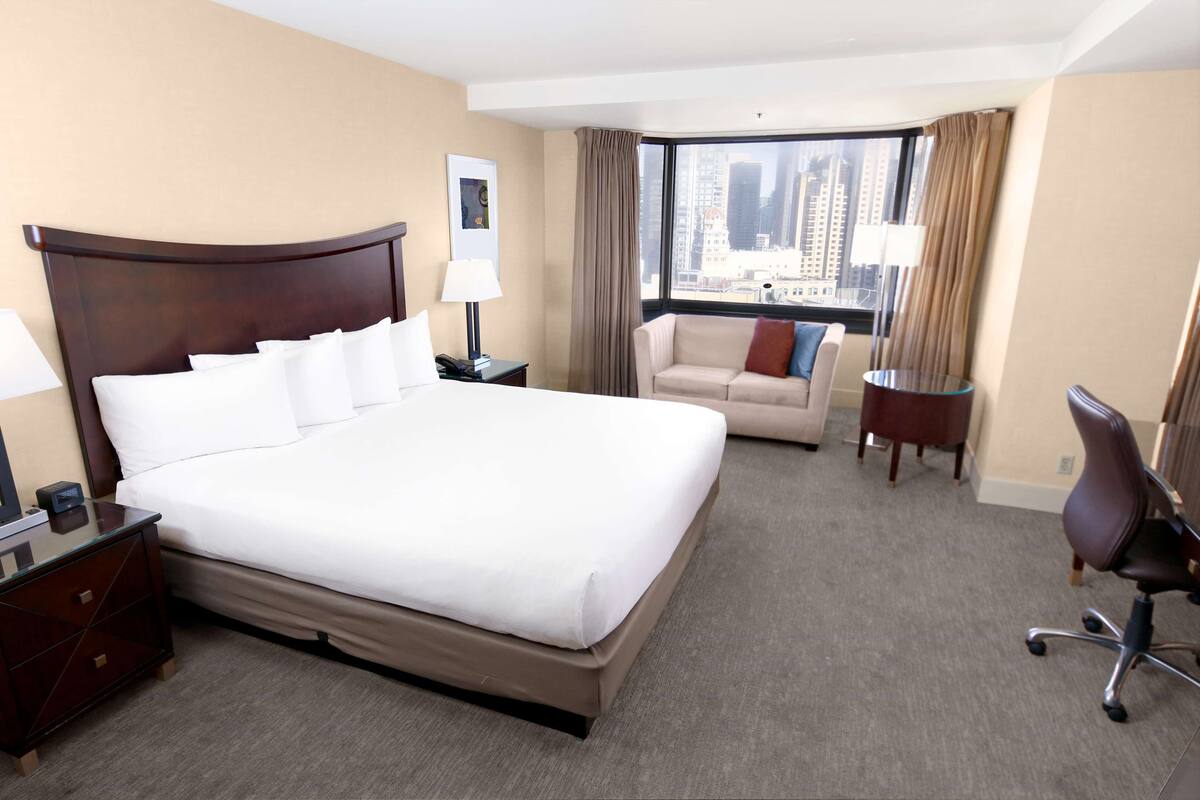 A hotel room with a large bed, a desk, a chair, and a view of the cityscape.