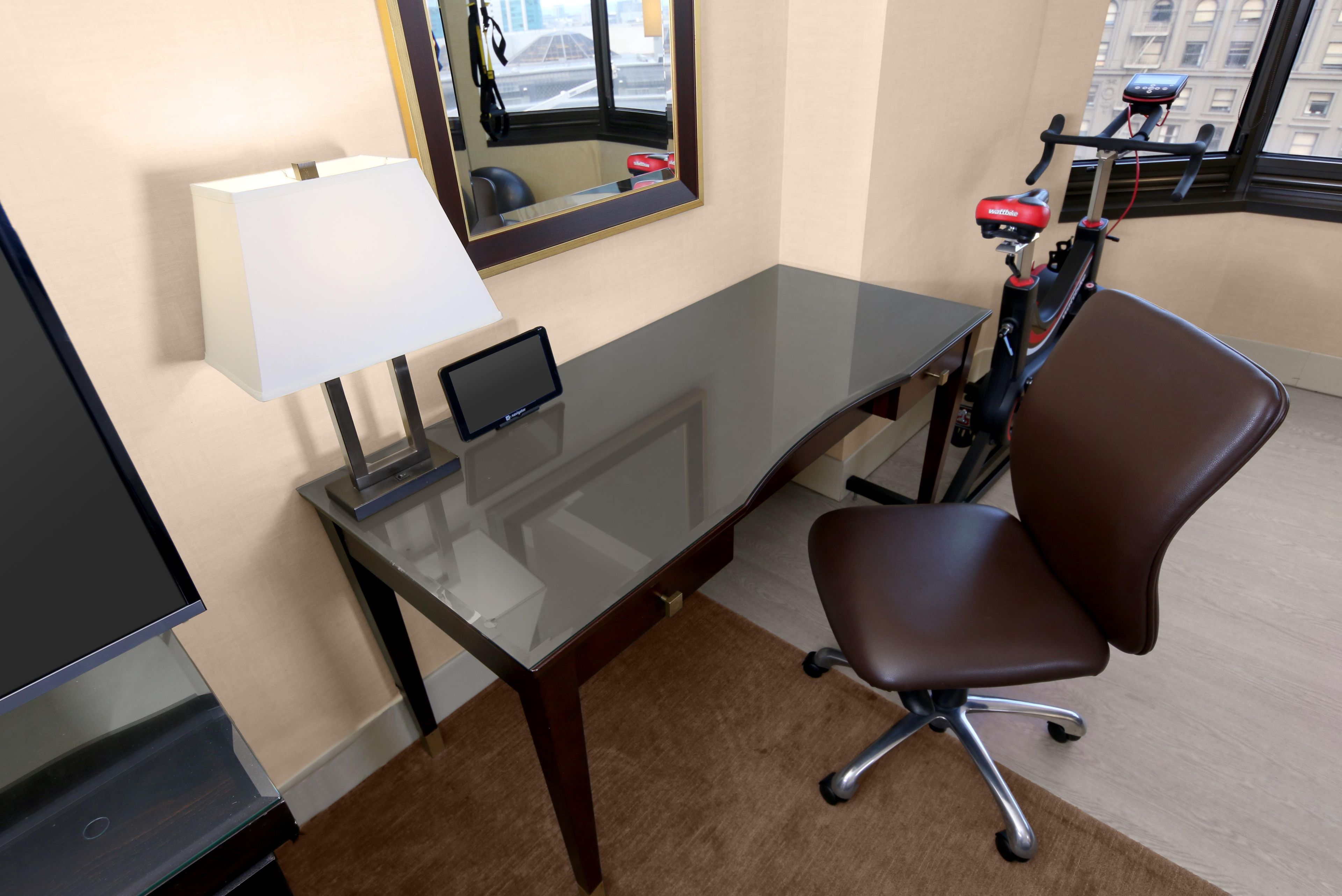 A hotel room with a desk, chair, lamp, and a treadmill.