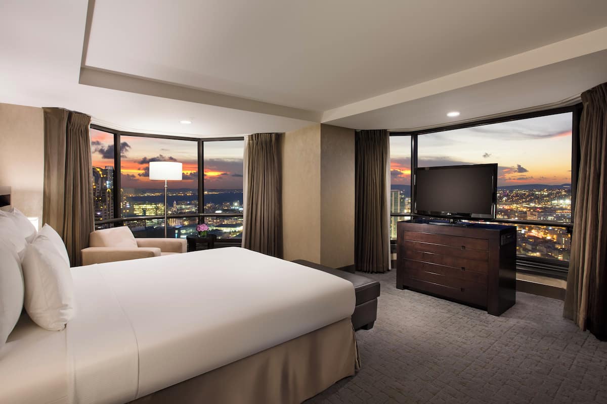 A hotel room with a large bed, a TV, a desk, and a view of a cityscape at sunset.