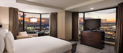 A hotel room with a large bed, a TV, a desk, and a view of a cityscape at sunset.