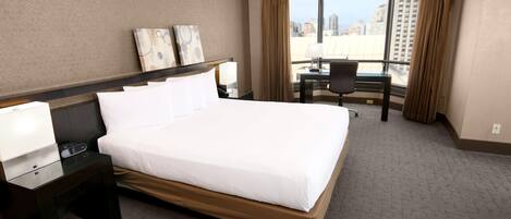 A hotel room with a large bed, bedside tables, a desk with a chair, and a view of the city through large windows.