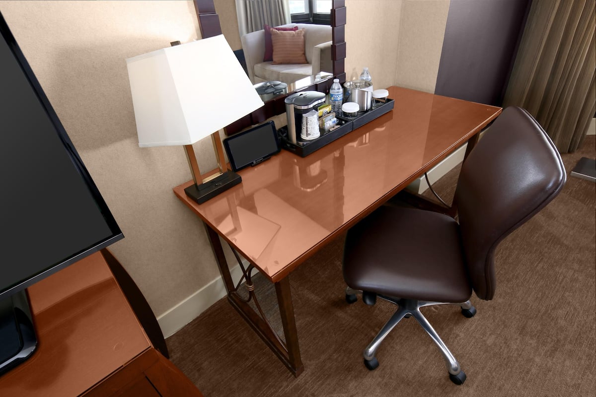 A hotel room with a desk, chair, lamp, and a tray with various items.