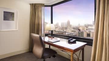 Habitación de hotel con escritorio, silla, lámpara y vistas a un paisaje urbano a través de un amplio ventanal.