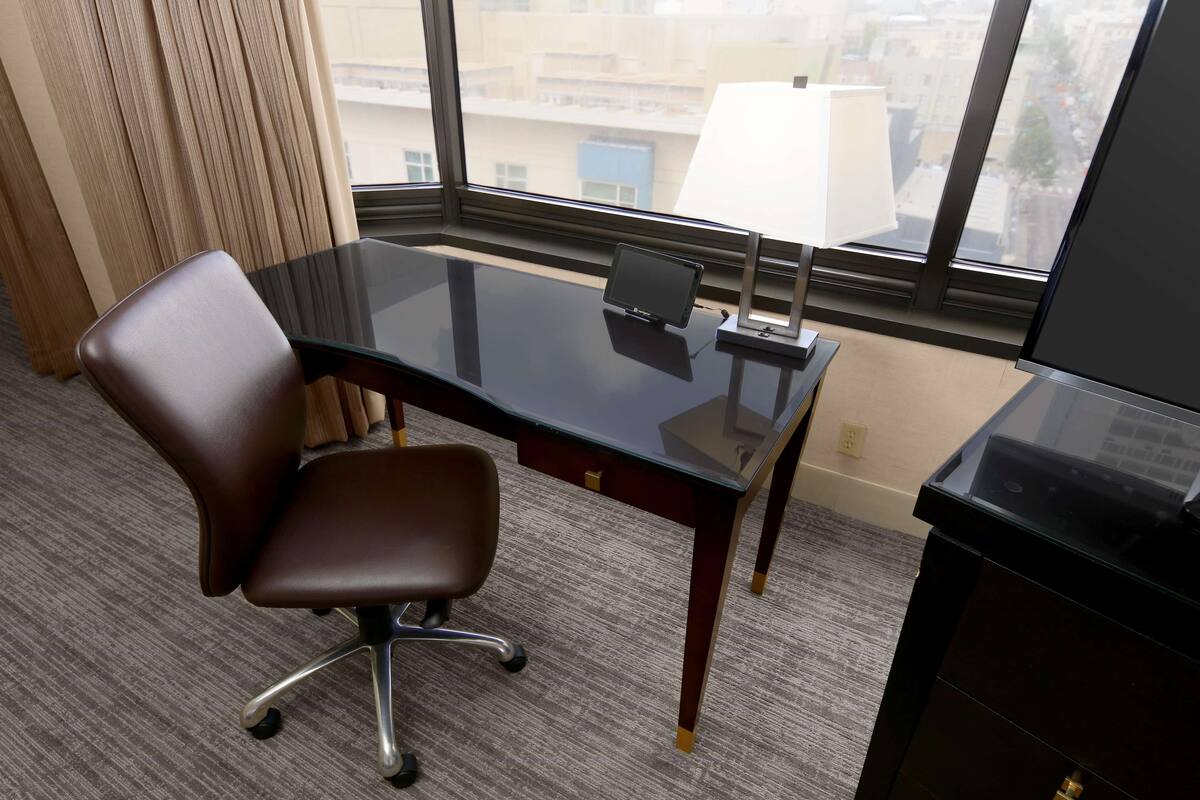 A hotel room with a glass desk, a brown office chair, a lamp, and a view of the city through large windows.