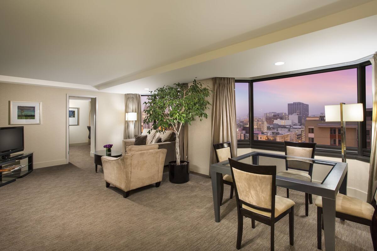 A hotel room with a city view, a sofa, a dining table with chairs, a TV, and a potted plant.