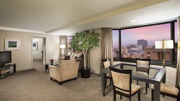 A hotel room with a city view, a sofa, a dining table with chairs, a TV, and a potted plant.