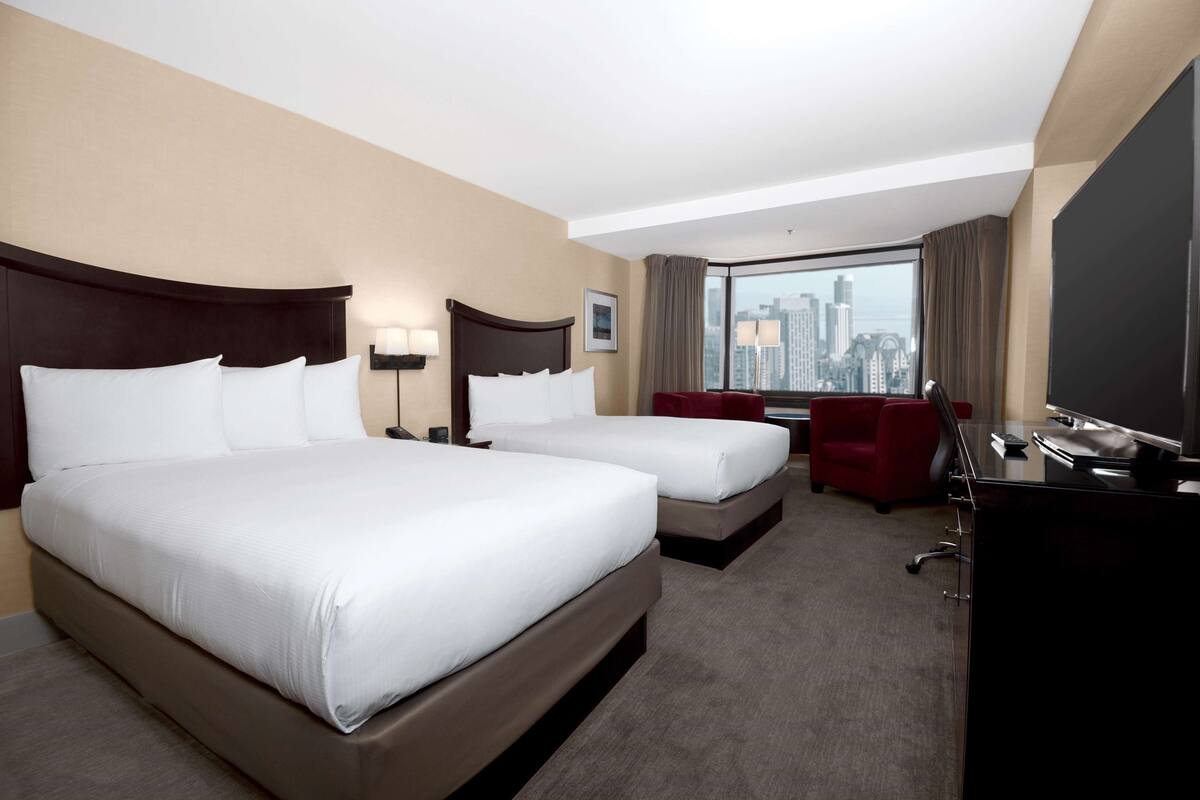 A hotel room with two beds, a desk, a chair, and a large window offering a city view.