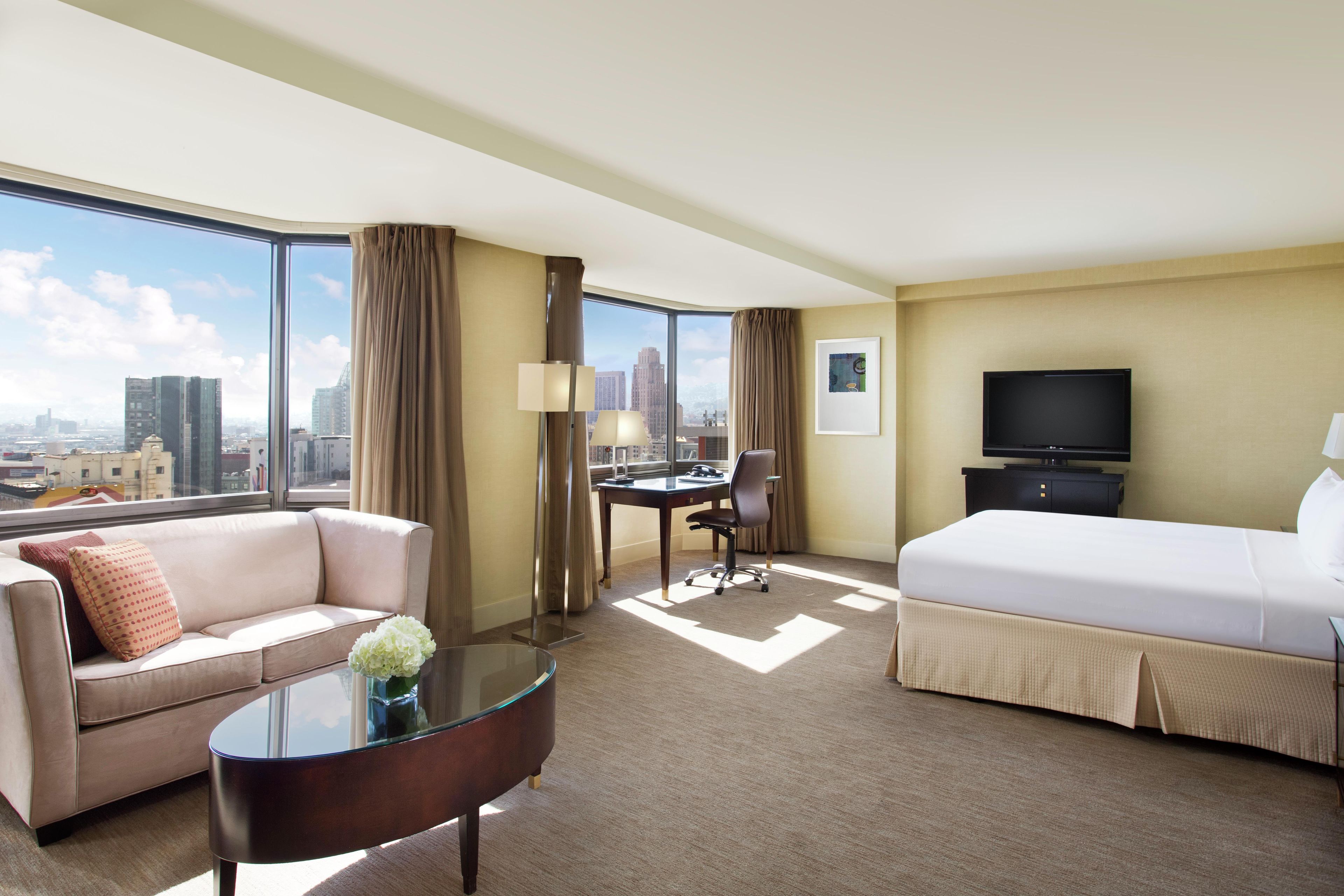 Habitación de hotel con cama, sofá, escritorio, televisor y vista a la ciudad.