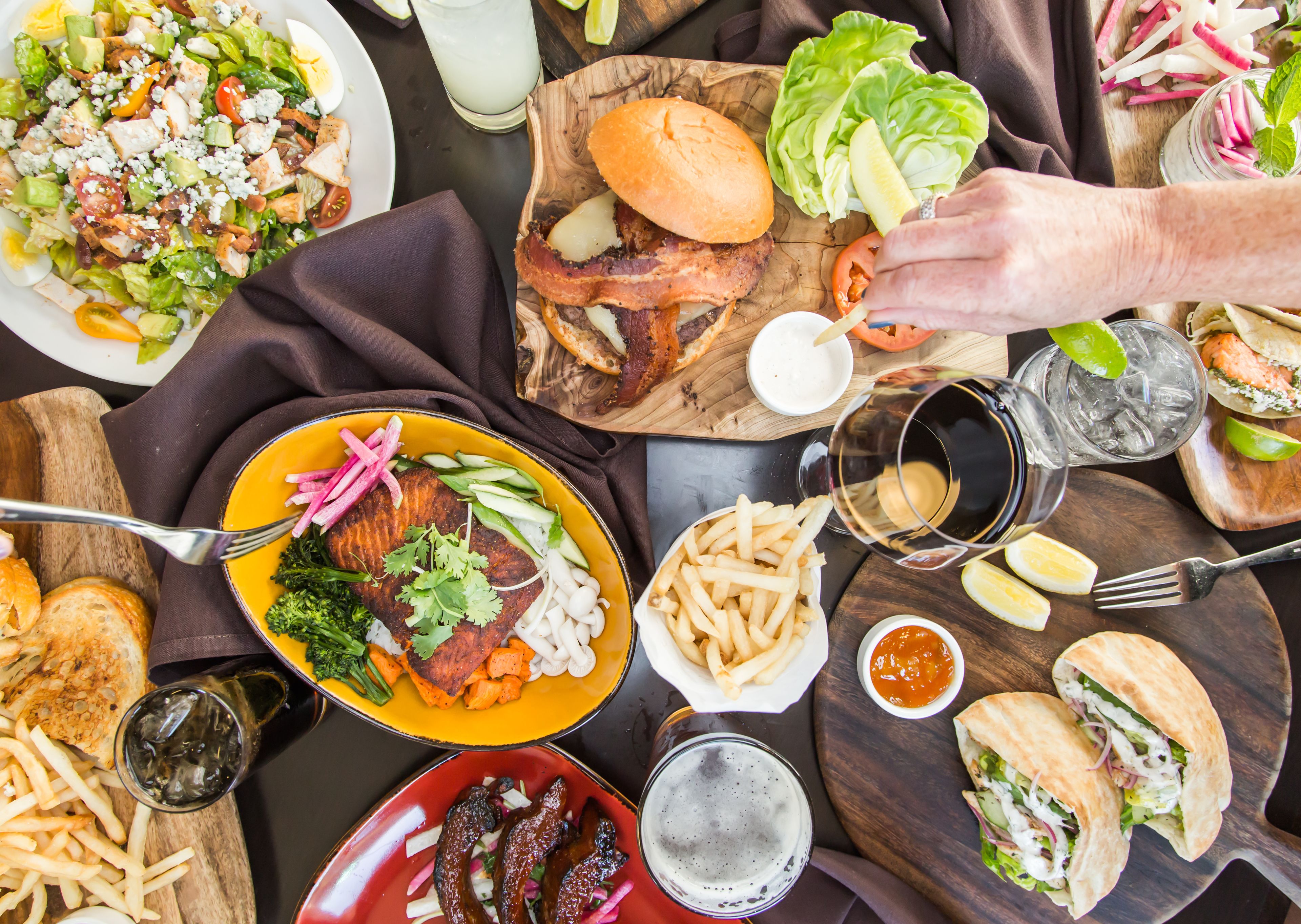 A variety of dishes including burgers, fries, and a salad, with a person pouring a drink.