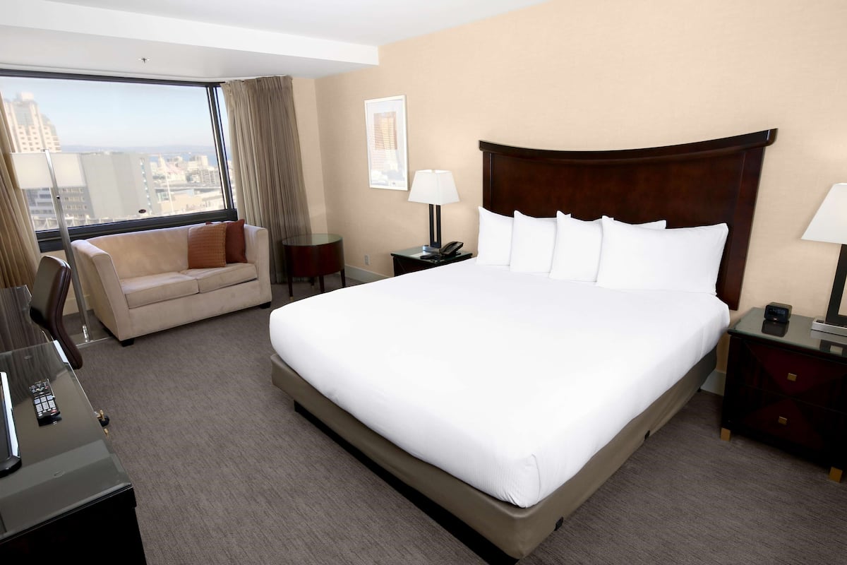 A hotel room with a large bed, a sofa, a desk with a phone, and a view of the city through a large window.