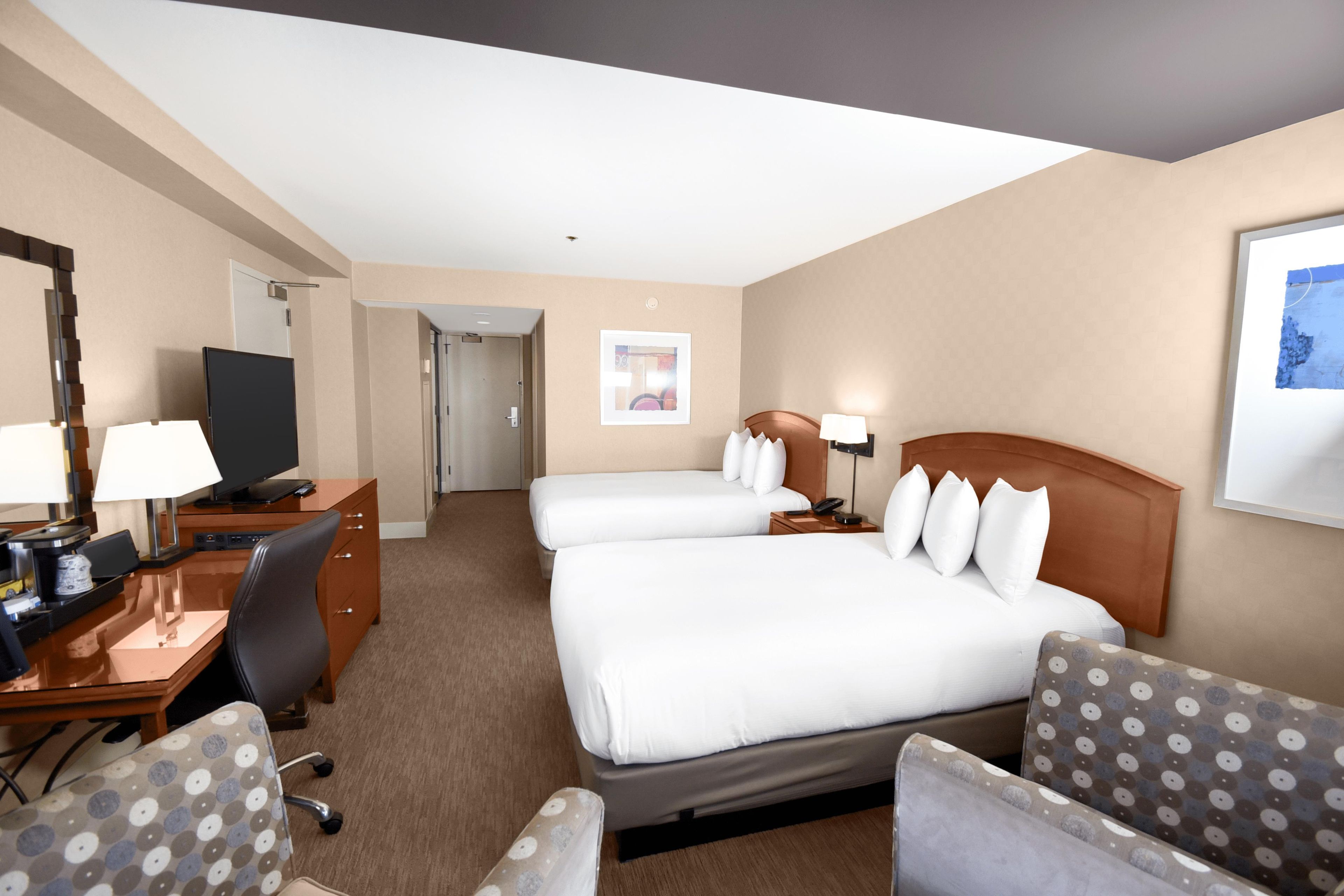 A hotel room with a desk, two beds, a chair, a television, and a framed picture on the wall.