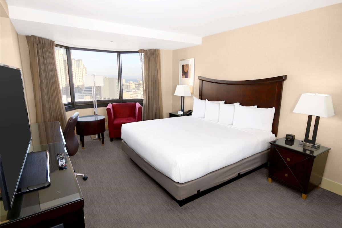 A hotel room with a large bed, a desk with a TV, a red chair, and a view of the city through the window.