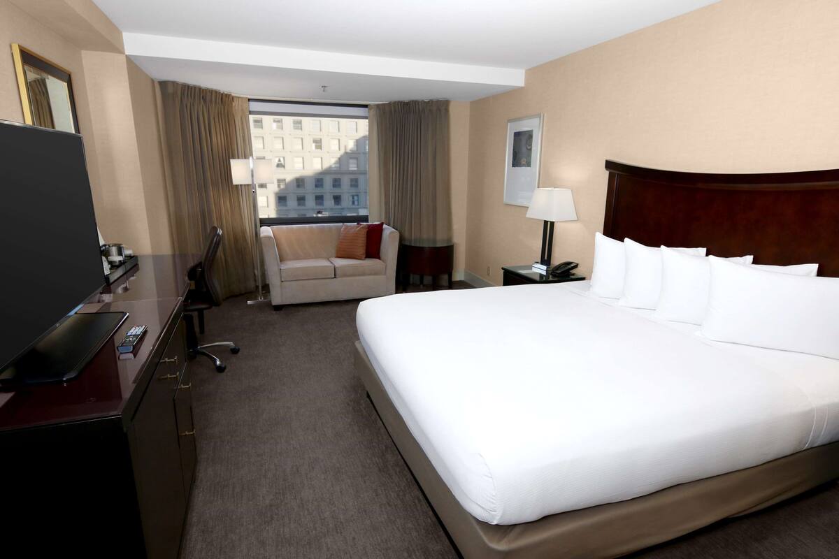 A hotel room with a large bed, a desk with a TV, a sofa, and a view of buildings through the window.