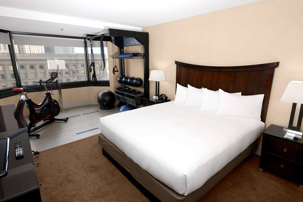 A hotel room with a large bed, exercise equipment, a desk, and a view of a cityscape.