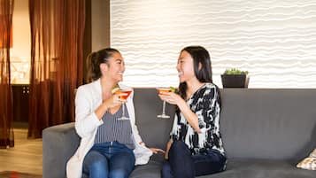 Two people sitting on a grey sofa, holding drinks, with a patterned wall and a plant in the background.