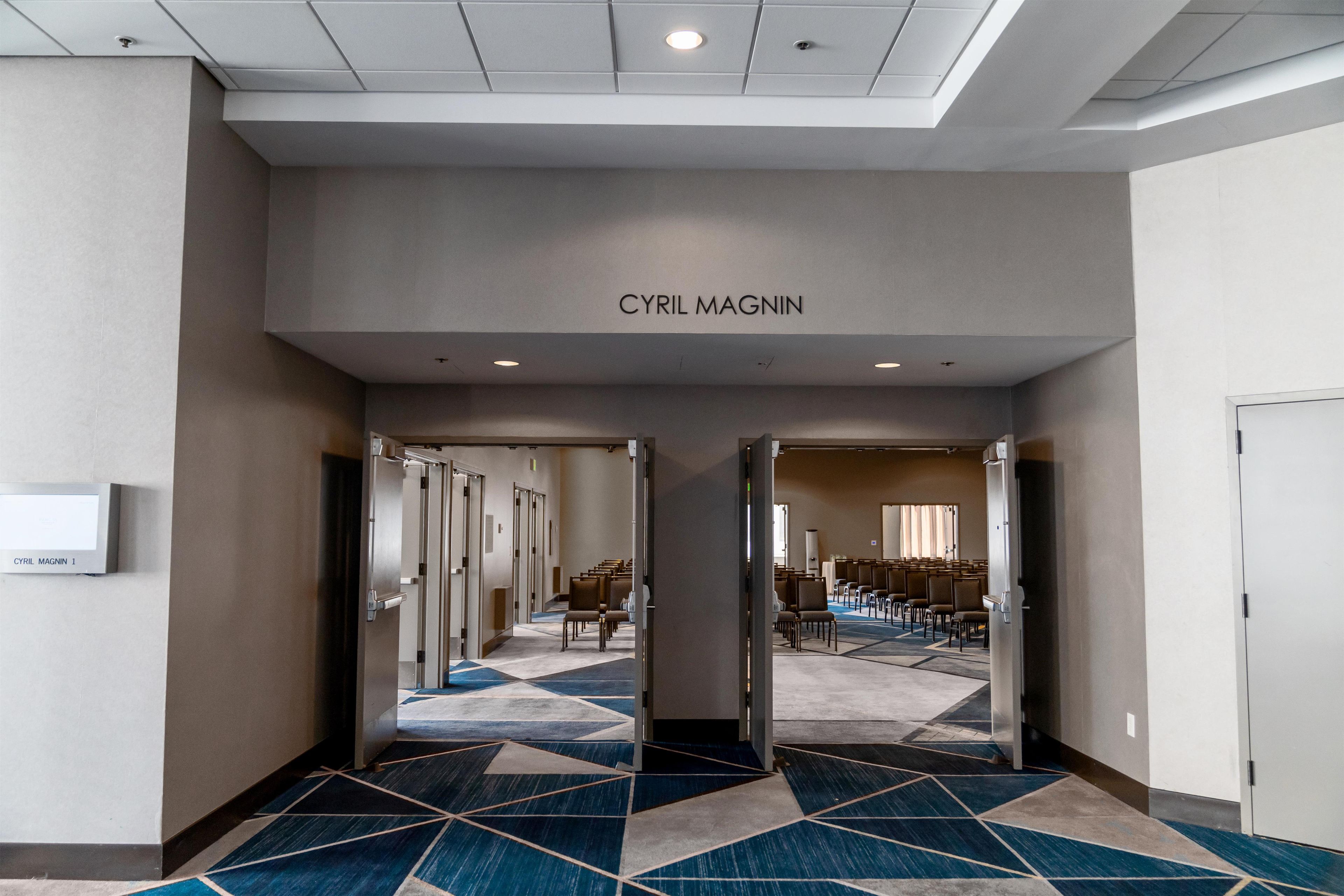 Et hotelbrugsgang med et geometrisk mønstret tæppe, døre der fører til et siddeområde, og et skilt med teksten 'CYRIL MAGNIN'.
