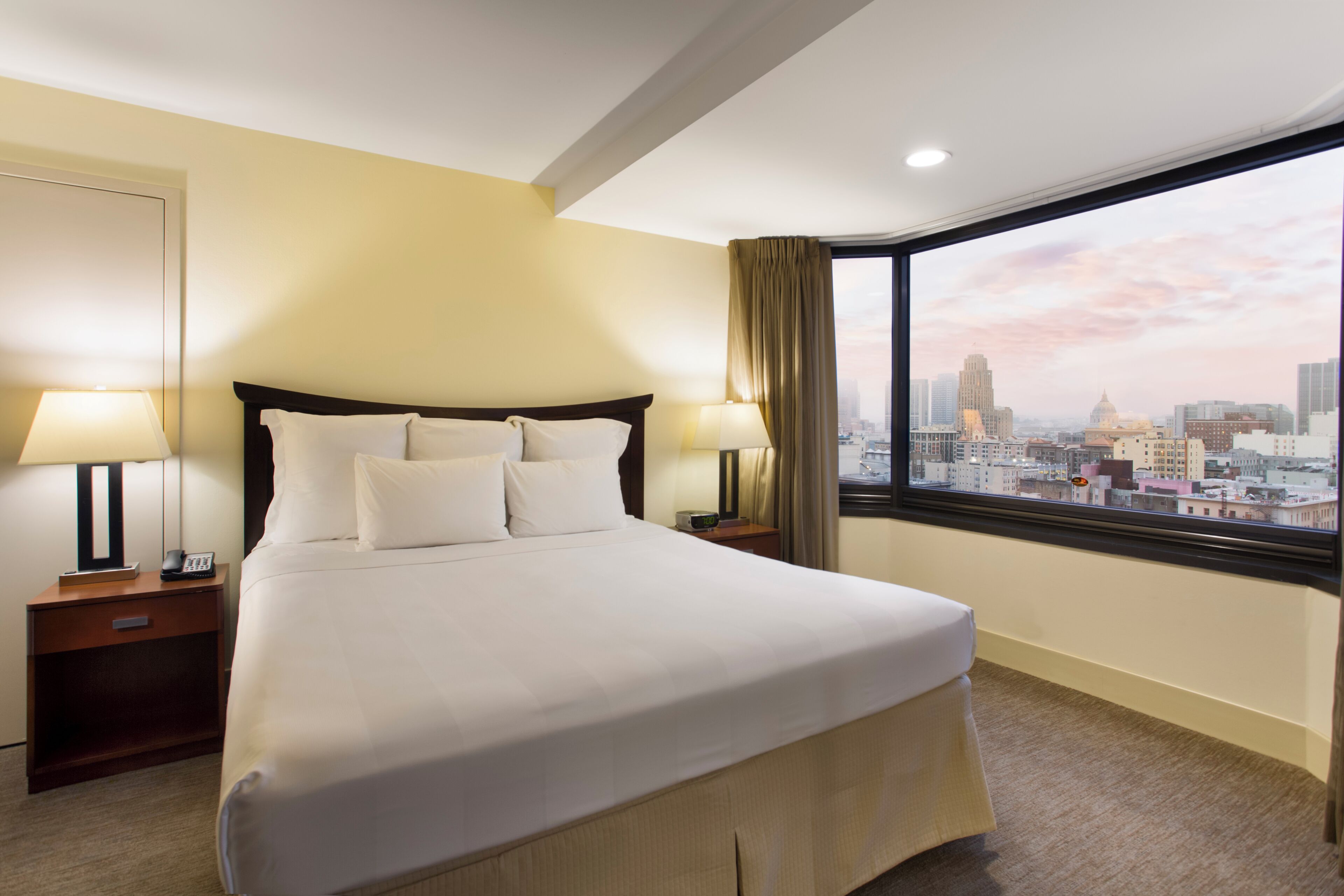 A hotel room with a large bed, two bedside tables with lamps, a view of a cityscape through a large window.