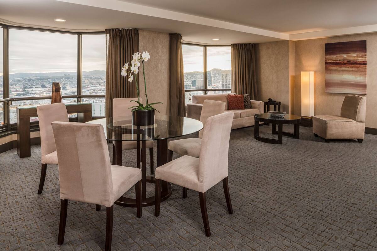 A modern hotel room with a large window offering a city view, a round glass table with chairs, a sofa, and a coffee table.