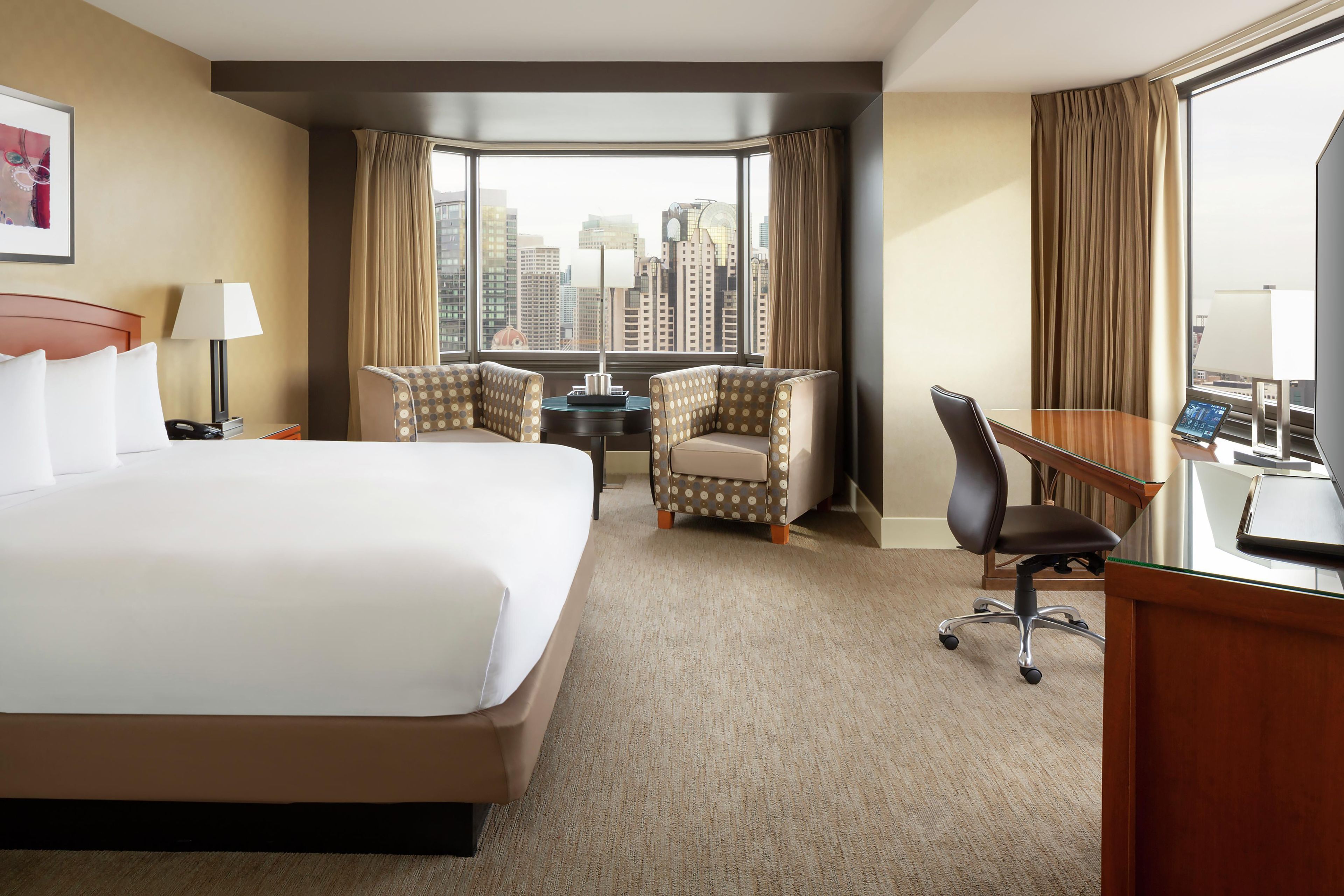 A hotel room with a large bed, two armchairs, a desk with a computer, and a view of the cityscape.