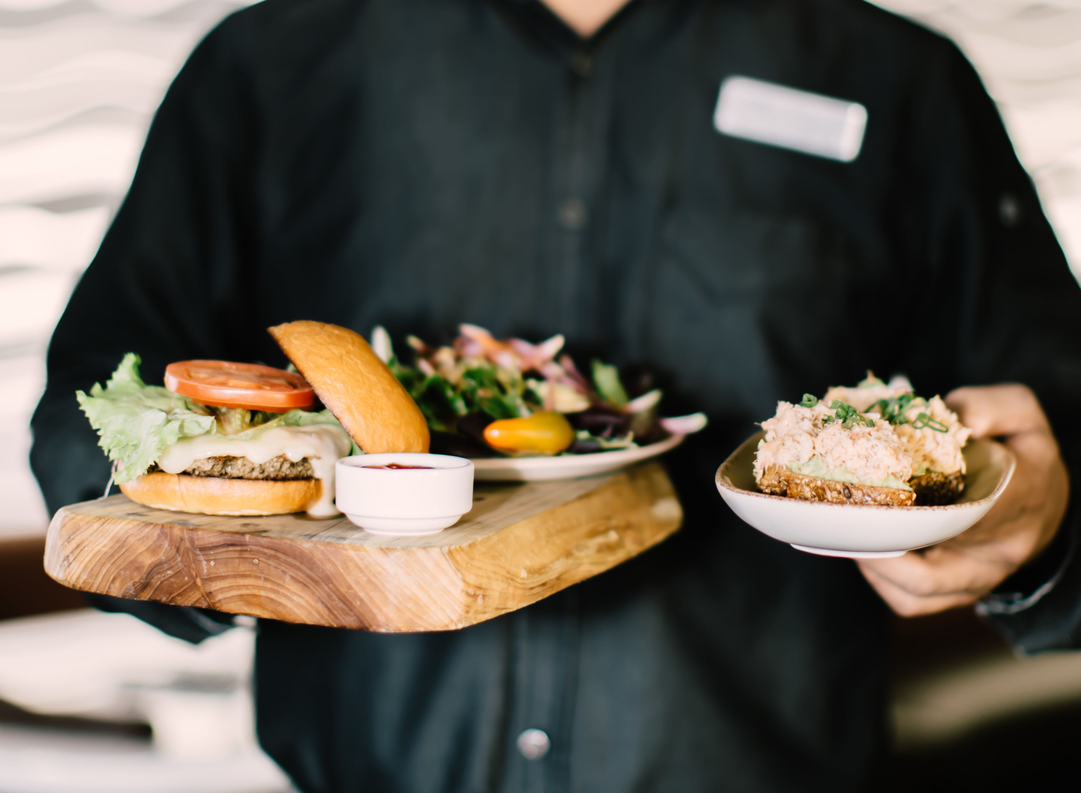 A person holding a wooden board with a burger, a salad, and a bowl of food.