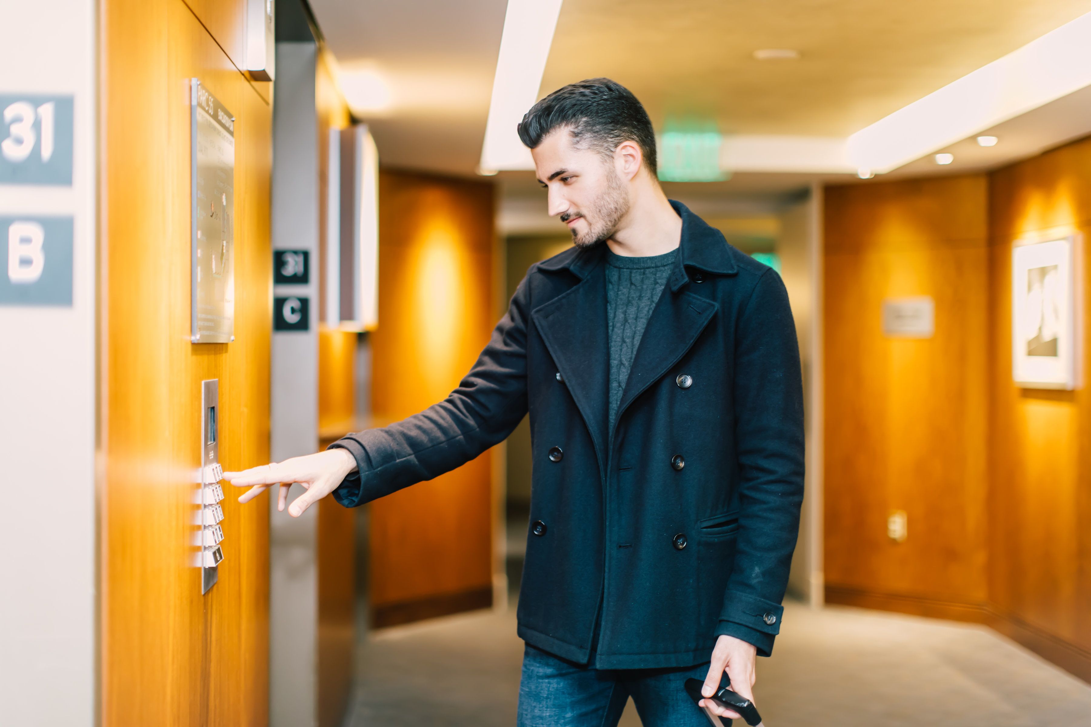 A man in a black coat interacting with an elevator control panel in a hotel hallway.