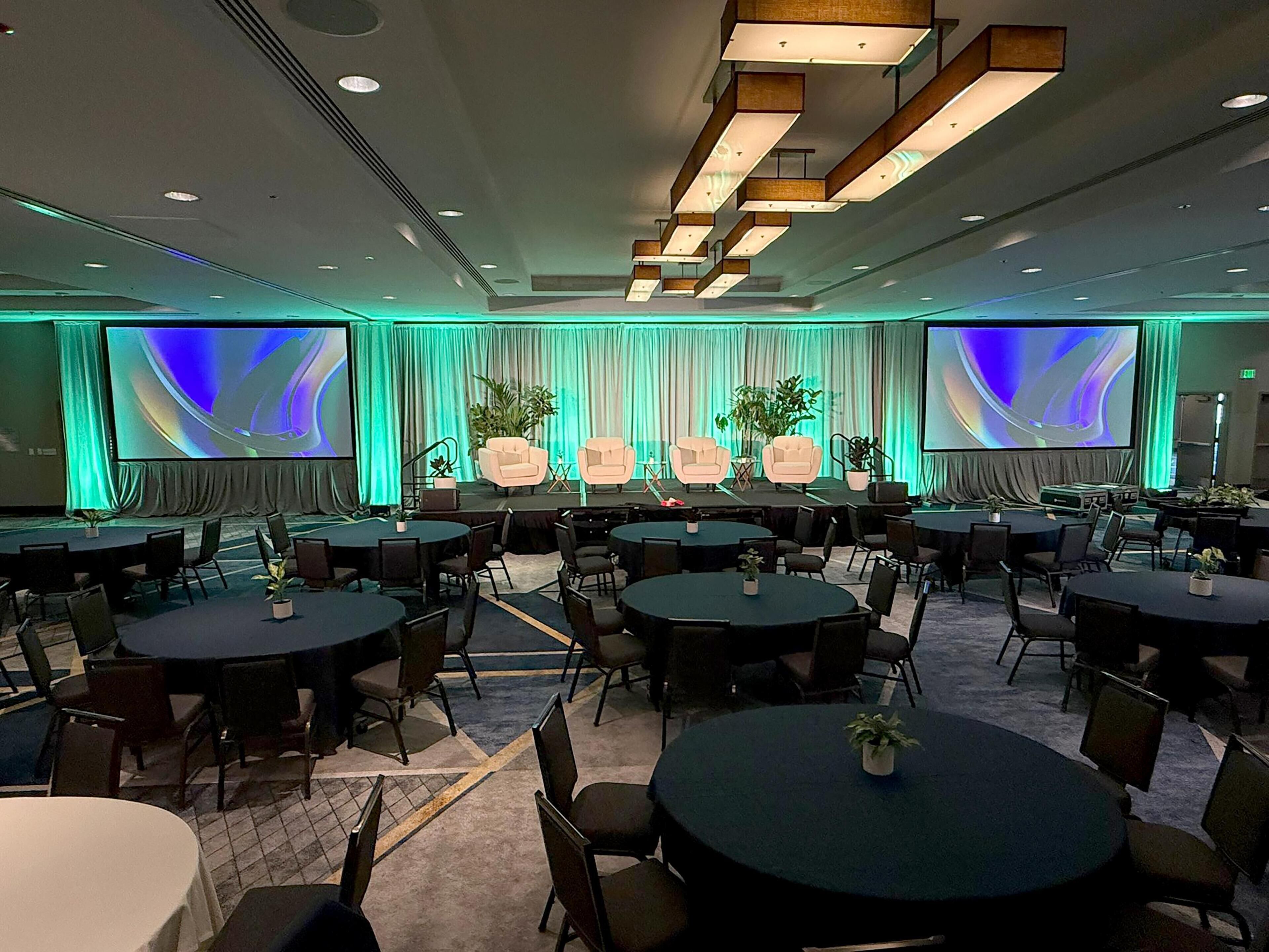 A conference room with multiple tables and chairs, a stage with a large screen, and green ambient lighting.