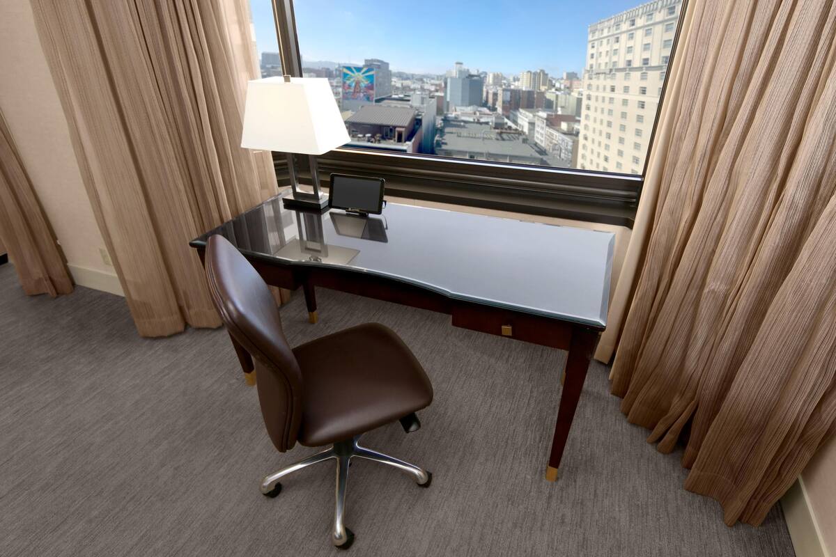 A hotel room with a desk, chair, lamp, and a view of the cityscape through the window.