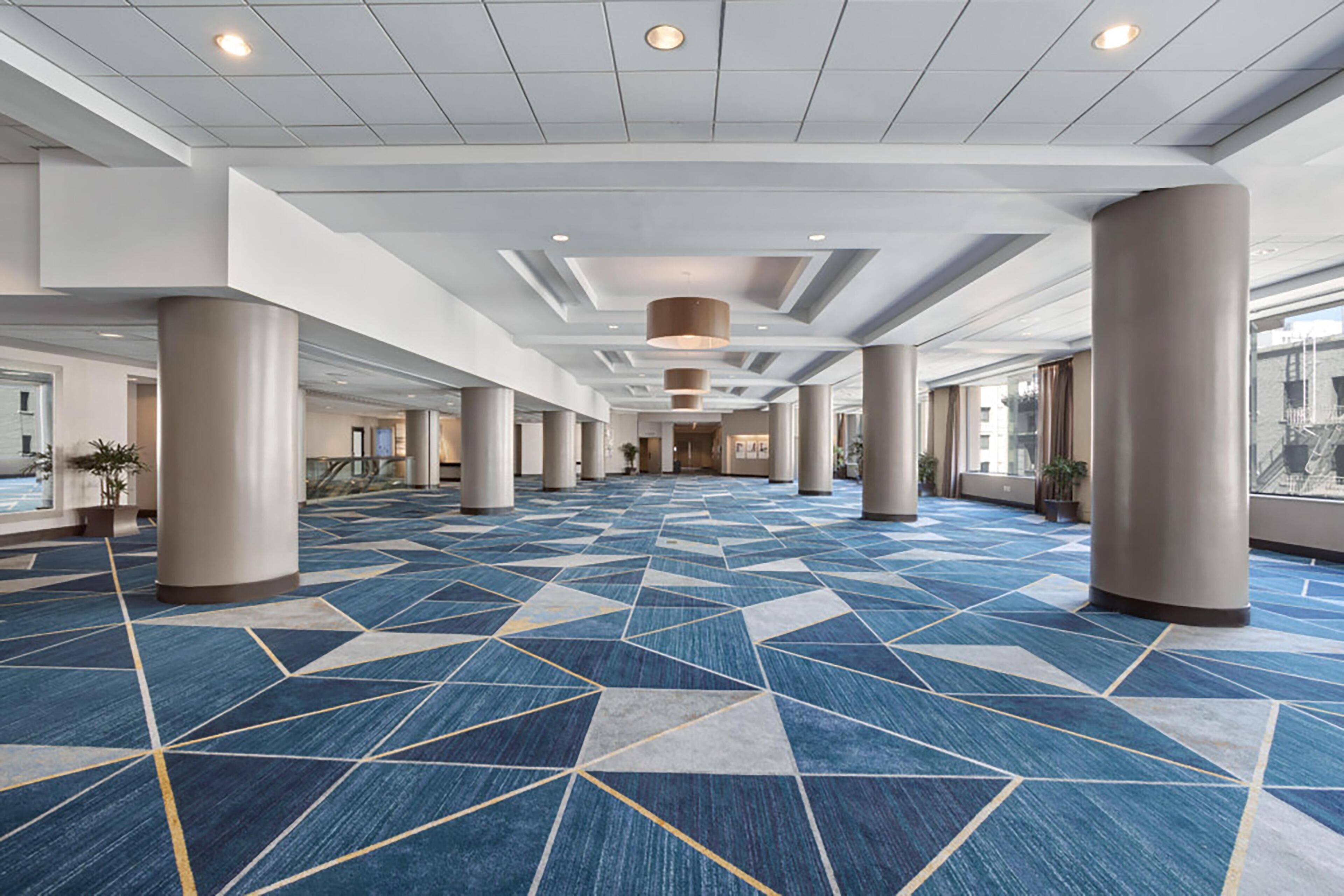 Un amplio vestíbulo de hotel con alfombra de diseño geométrico, grandes columnas y un techo alto con iluminación empotrada.