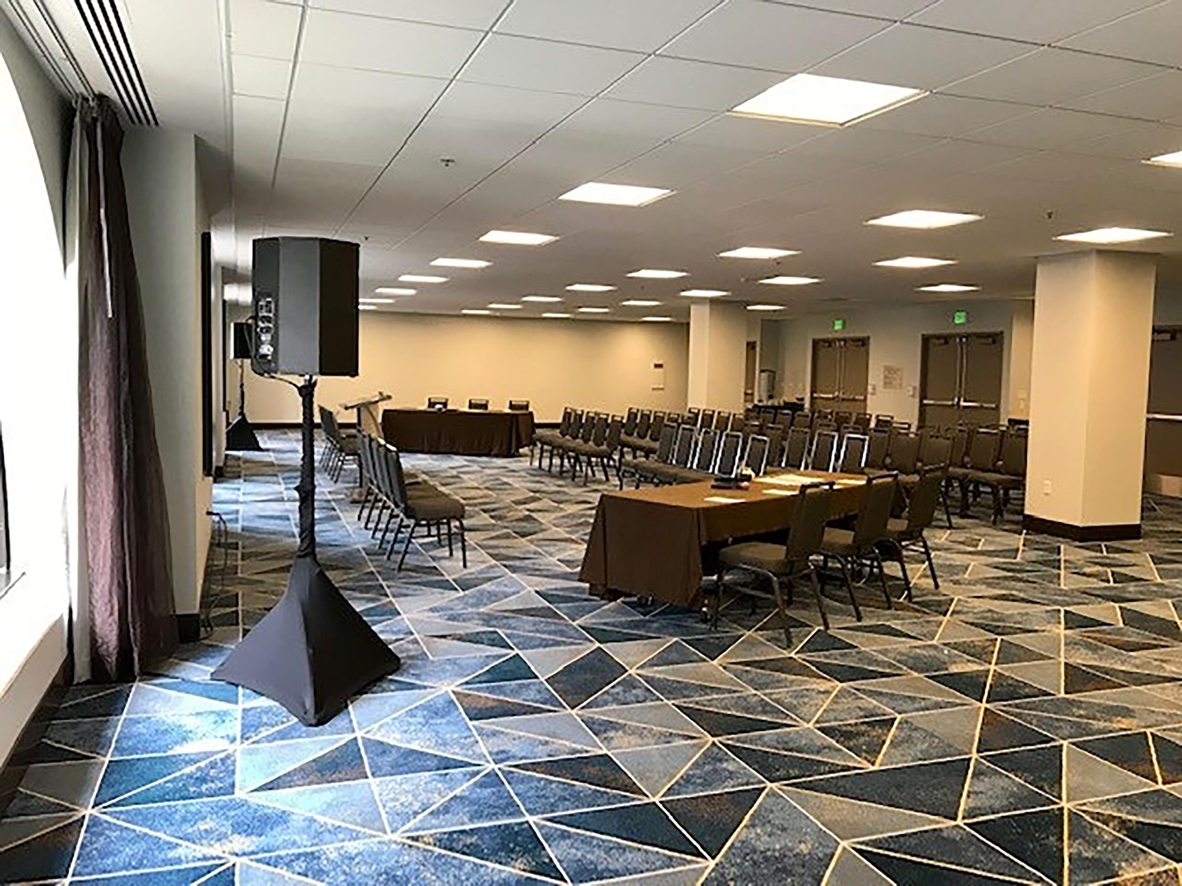 Un salón de conferencias con suelo de diseño geométrico, filas de sillas, una mesa central y un orador.