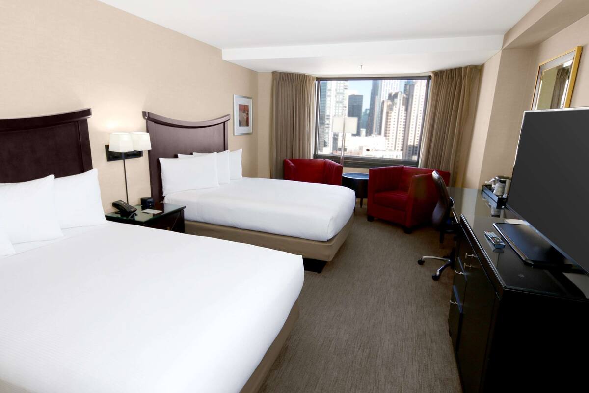 A hotel room with two beds, a desk, a chair, a TV, and a window with a city view.