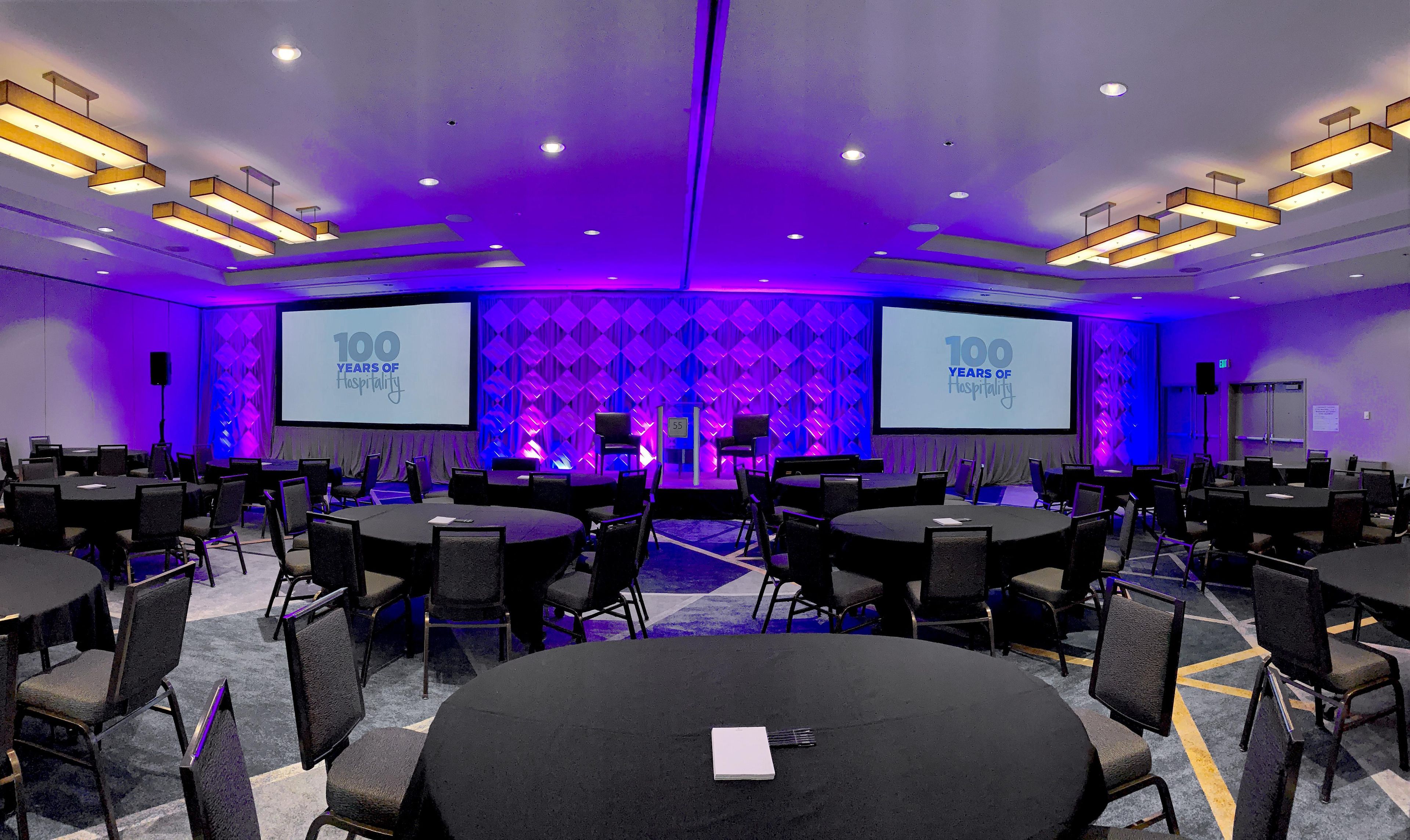 A conference room with multiple tables and chairs, a large screen displaying '100 YEARS OF Hospitality,' and a stage with a podium.