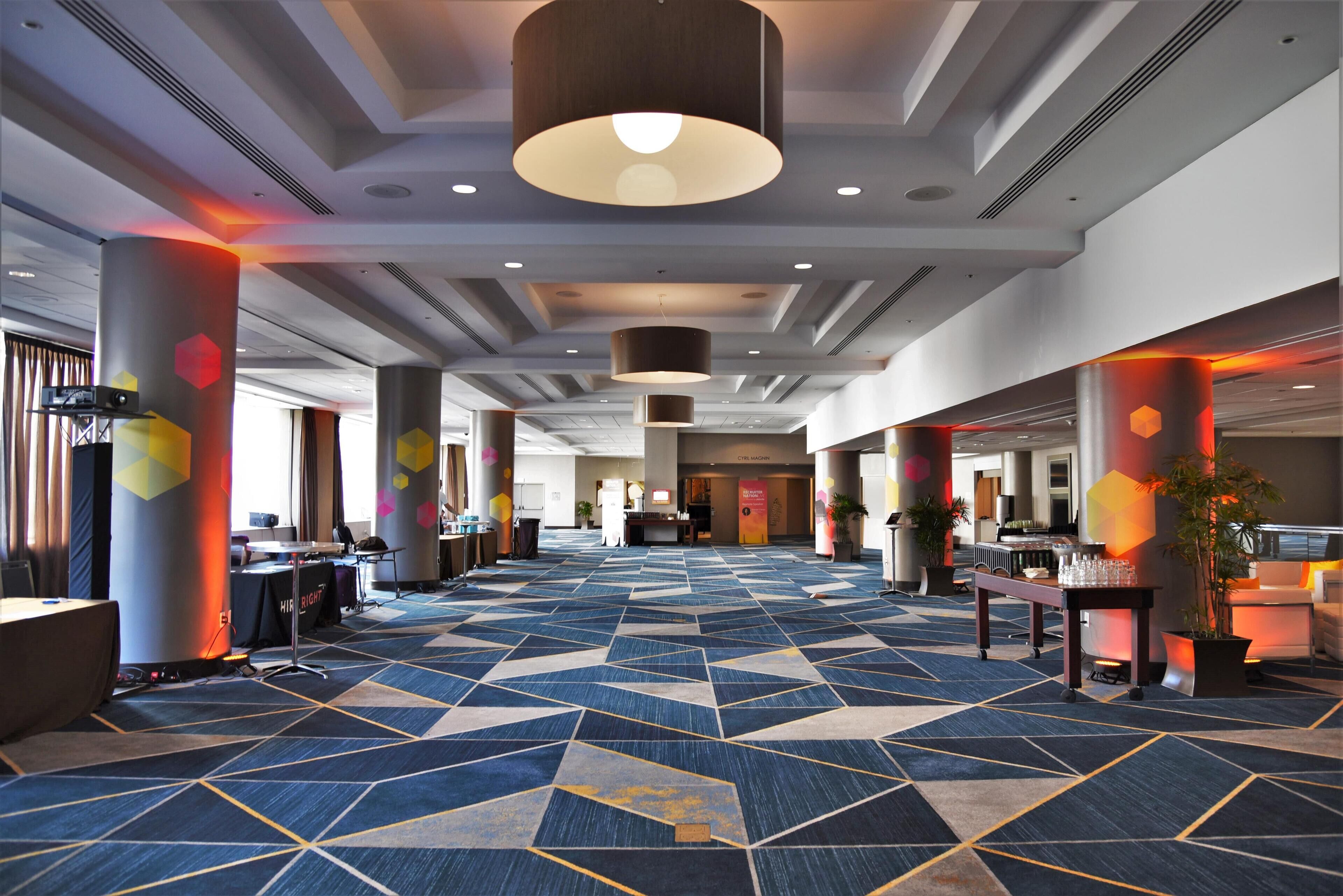 A spacious hotel lobby with a geometric patterned carpet, multiple seating areas, and modern lighting fixtures.
