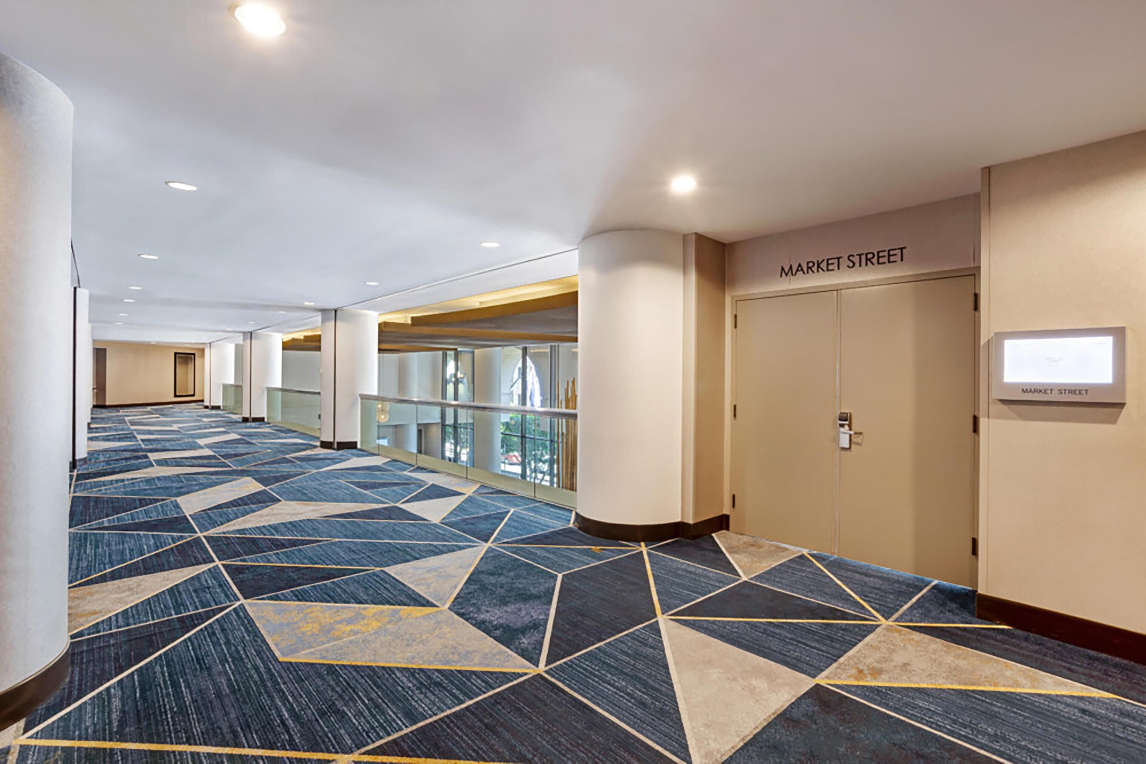 A hotel corridor with a geometric patterned carpet, a door labeled 'MARKET STREET', and a glass railing in the background.