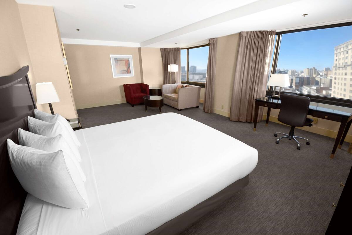 A hotel room with a large bed, a desk, a red armchair, and a view of the city.