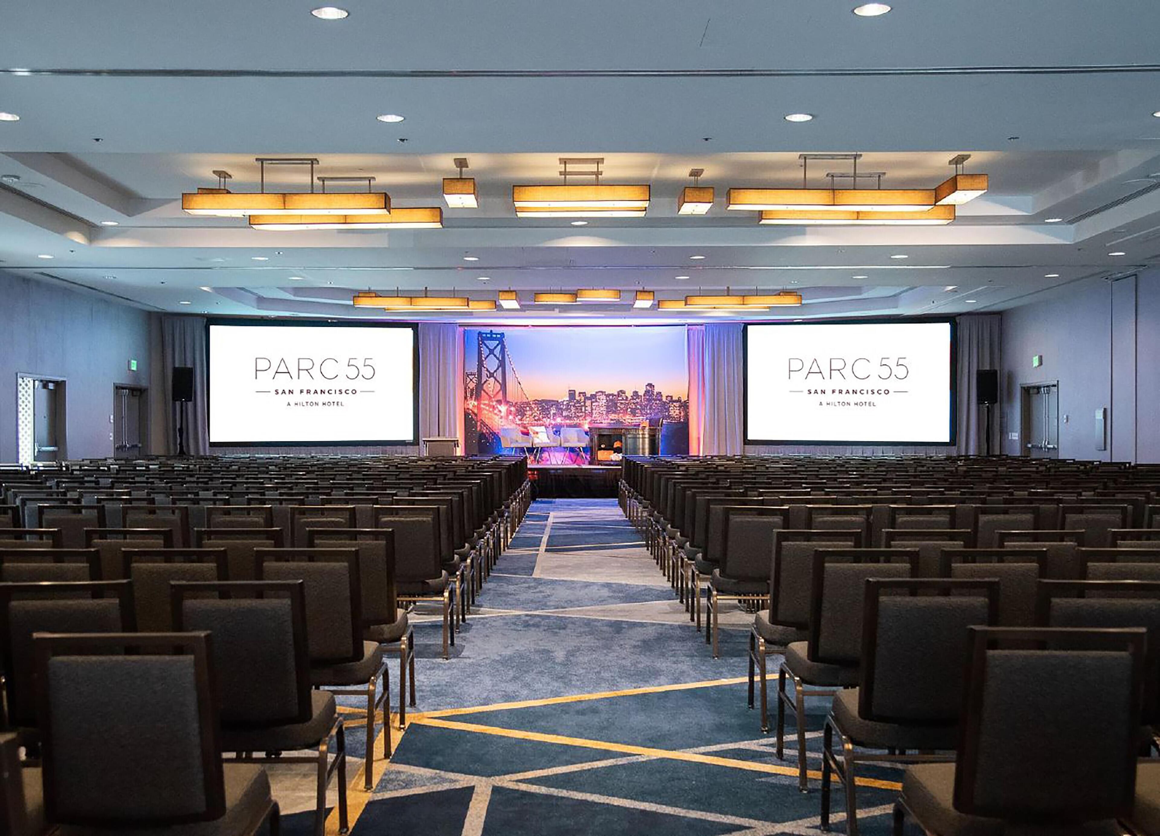 Ett konferensrum med rader av stolar, en stor skärm som visar texten ”PARC 55 SAN FRANCISCO, ETT HILTON-HOTEL” och en stadsbild i bakgrunden.