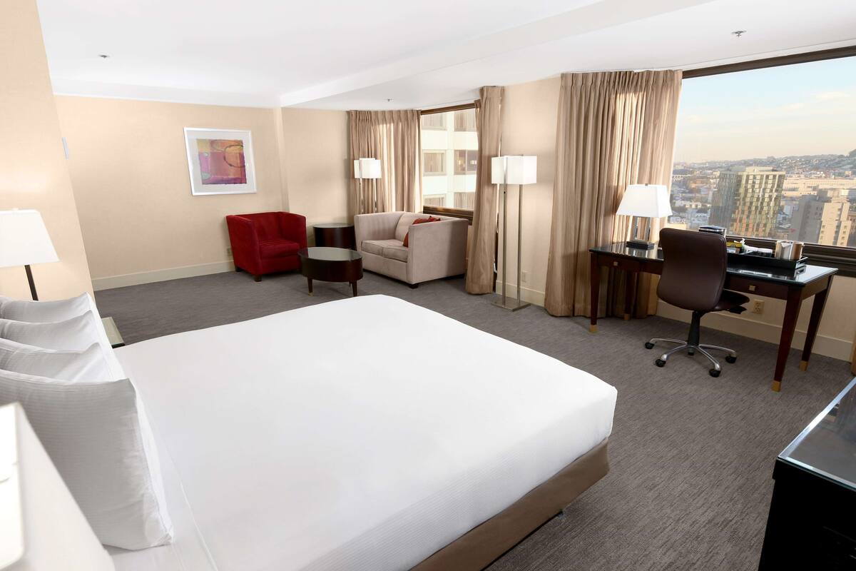 A hotel room with a large bed, a desk, a red chair, a sofa, and a view of the city.