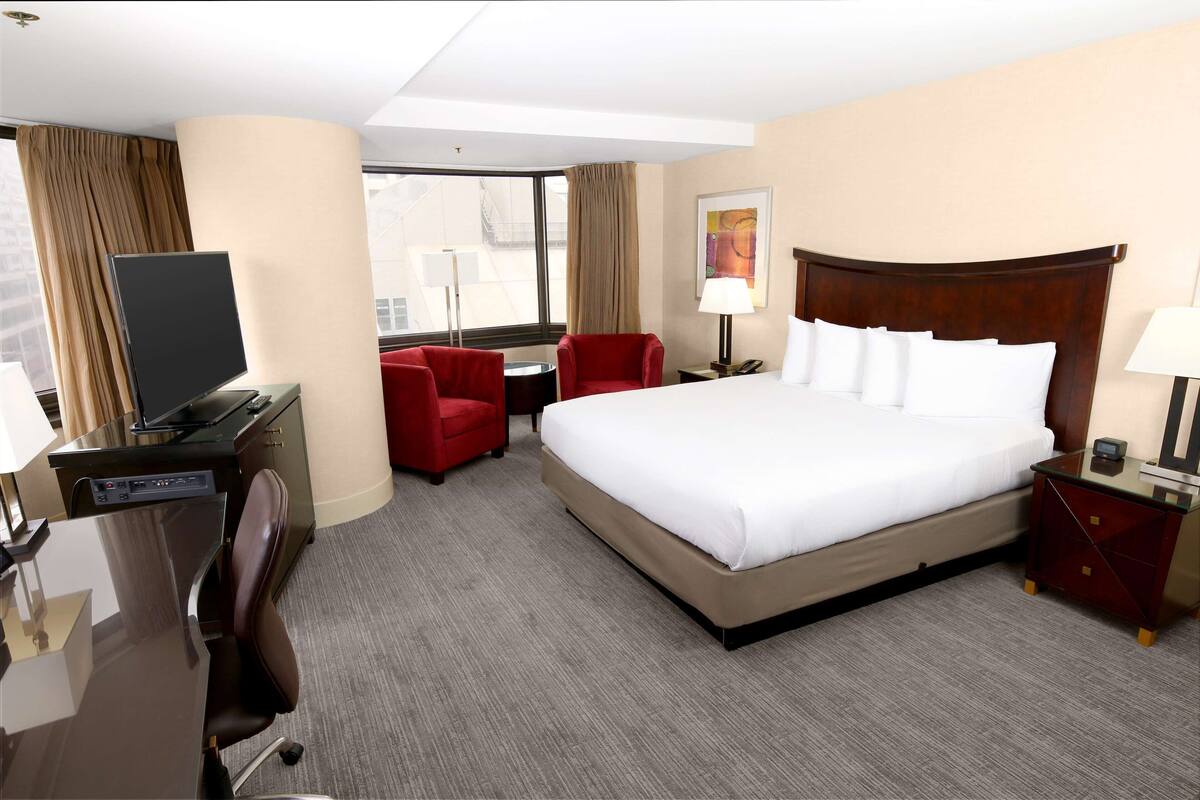 A hotel room with a large bed, a desk with a computer, a red chair, and a TV.