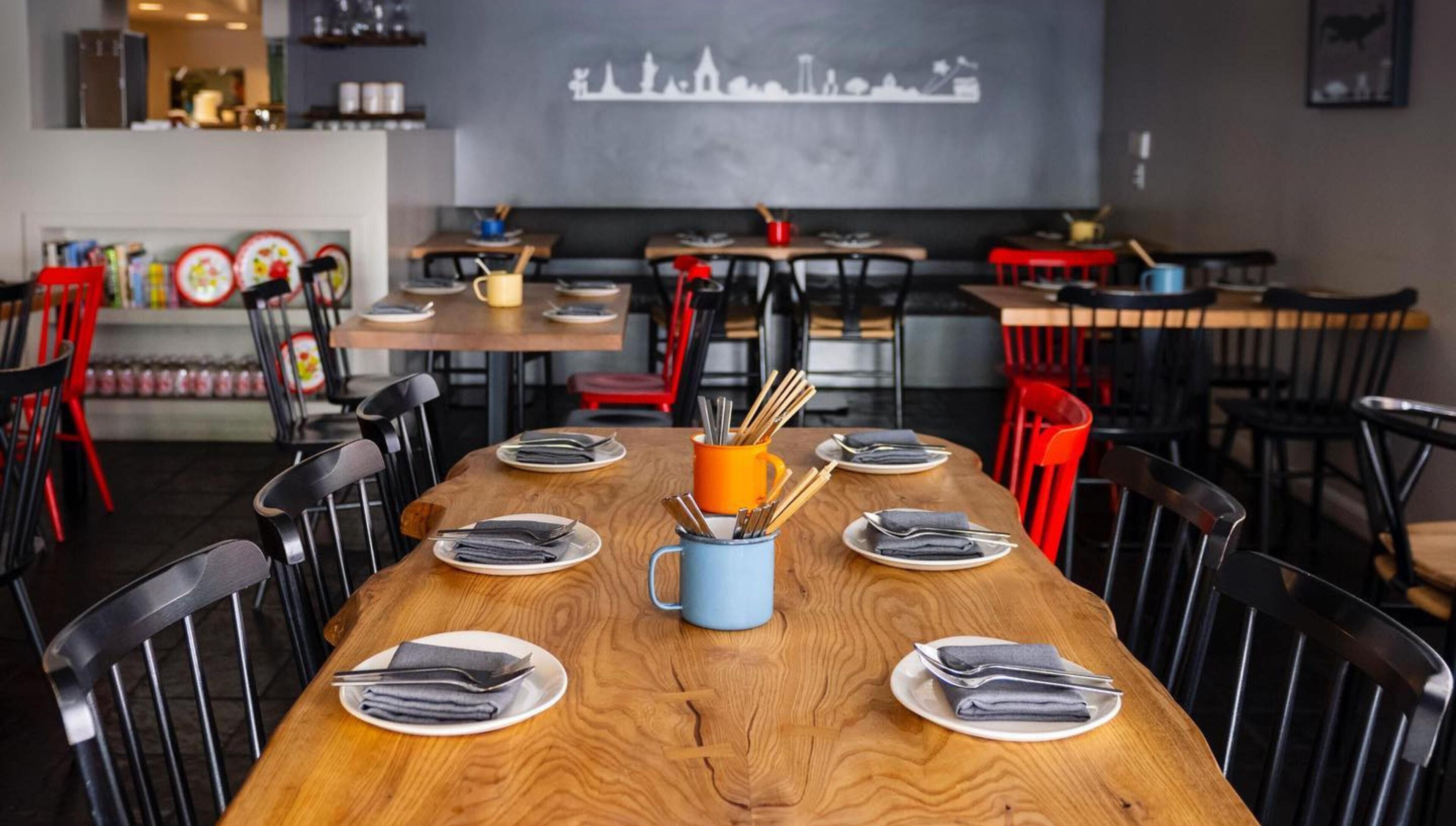 Ein Restaurantinneres mit einem langen Holztisch, gedeckten Plätzen, schwarzen Stühlen und einer Wandmalerei mit Stadtansicht.