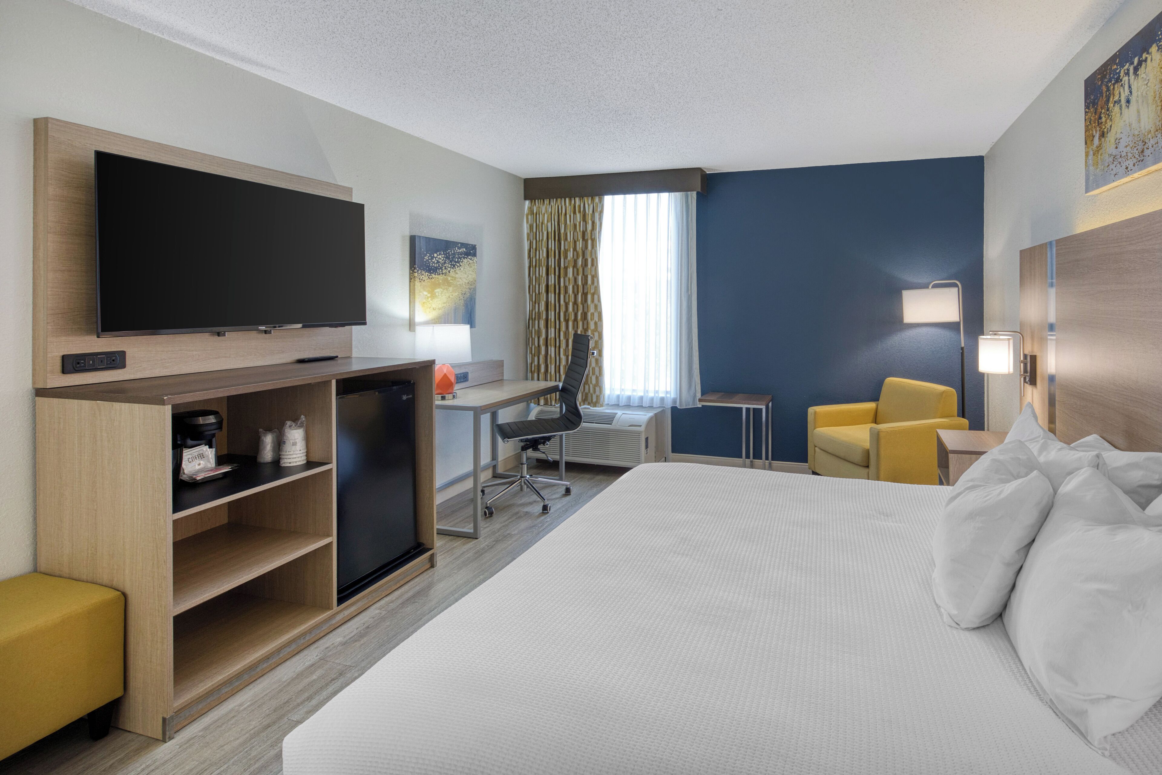 A hotel room with a large bed, a desk with a computer, a TV, and a yellow armchair.