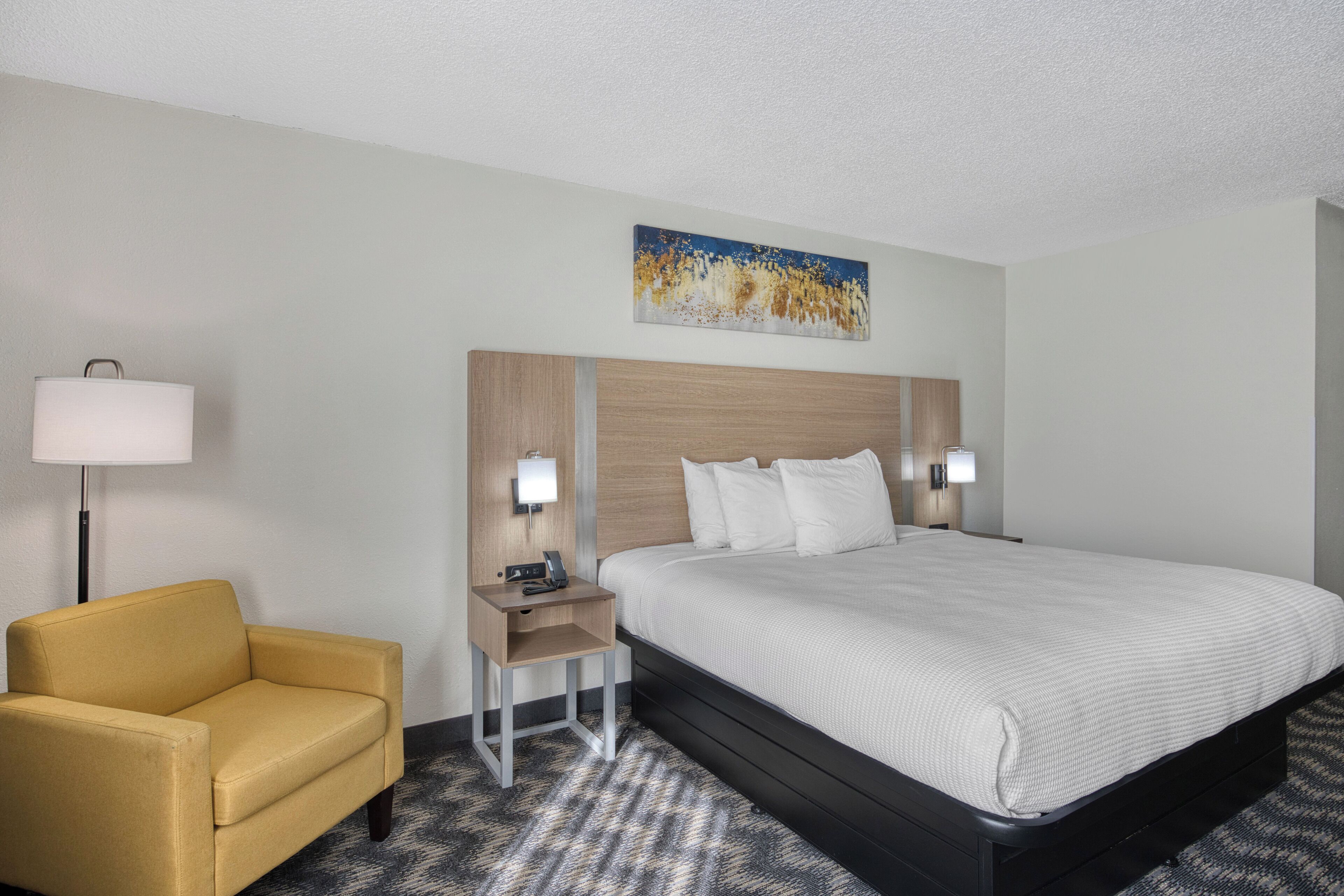 Una habitación de hotel con cama, mesita de noche, lámpara, una silla y un cuadro en la pared.