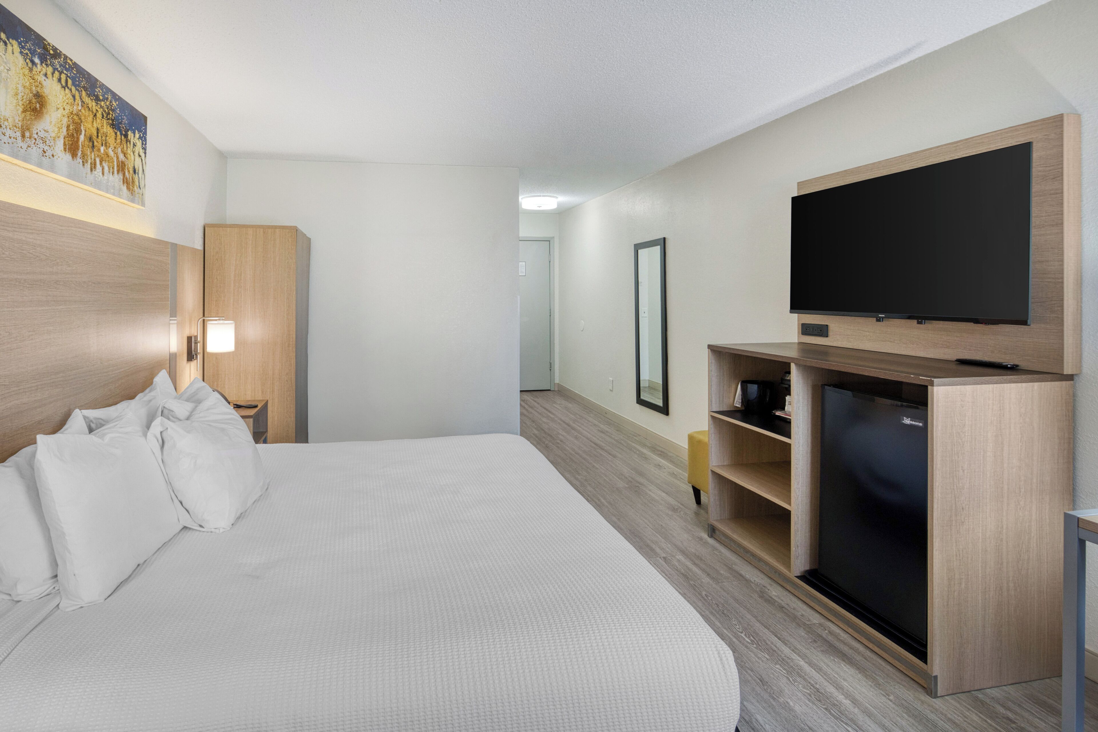 A hotel room with a large bed, a flat-screen TV mounted on a wooden cabinet, a bedside table with a lamp, and a painting on the wall.