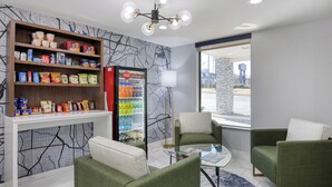 A modern hotel lobby with green upholstered chairs, a glass coffee table, a Coca-Cola refrigerator stocked with beverages, and a cityscape view through a large window. - Best Western Greensboro Coliseum Inn (Greensboro)