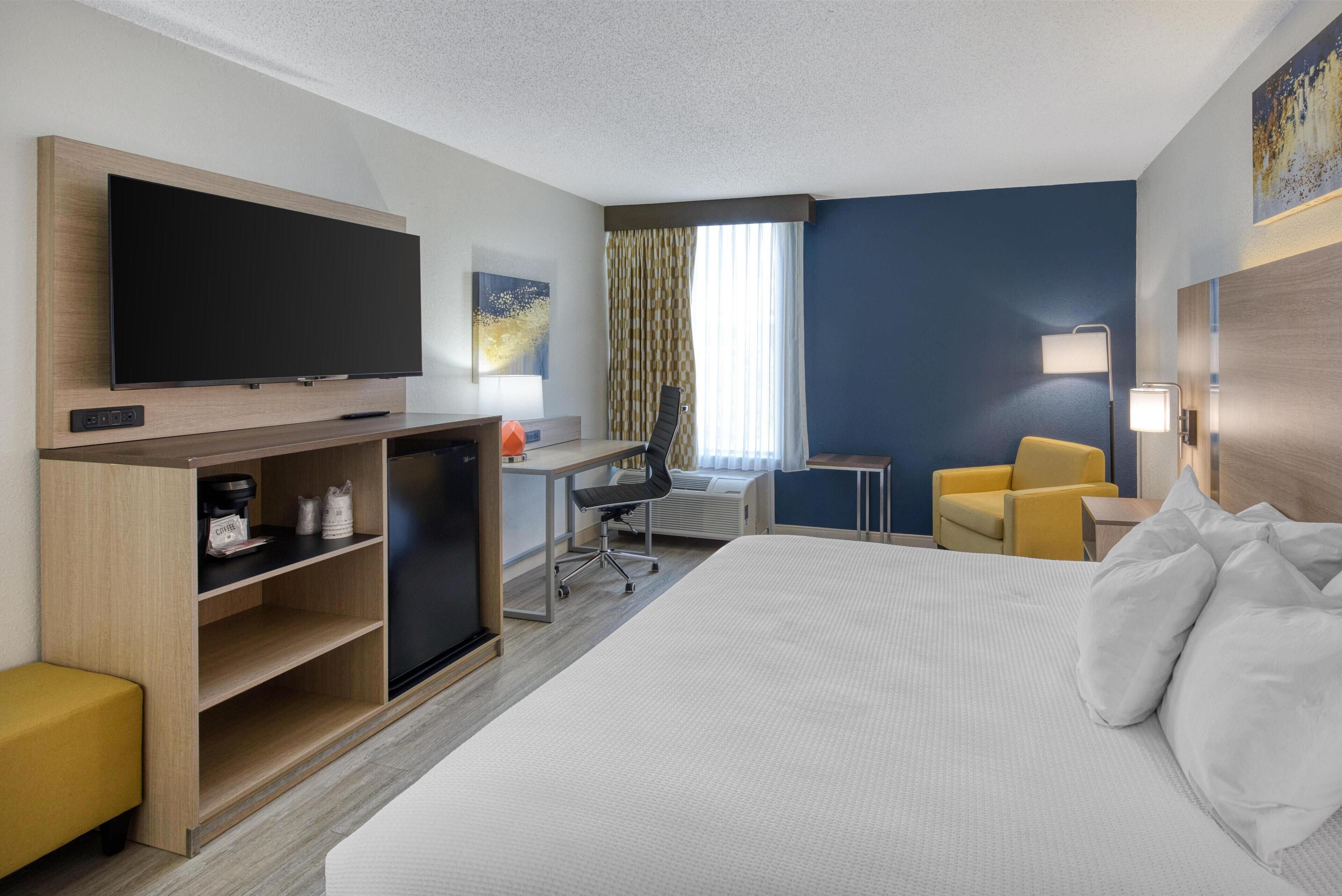 A hotel room with a large bed, a desk with a computer, a television, a coffee machine, and a yellow armchair.